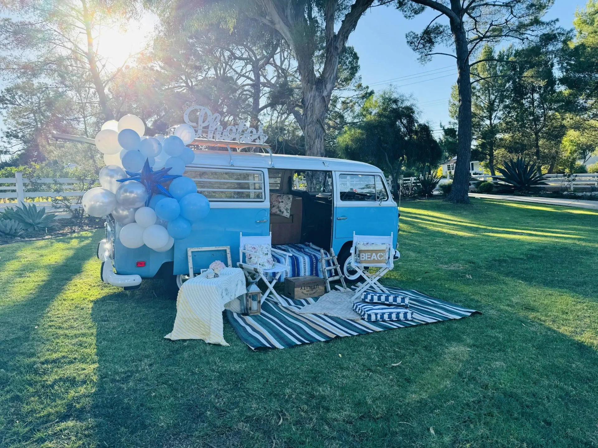 Sky blue 1970 VW Photobus on a sunlit lawn with a Photos marquee sign, blue and white balloon arch and a coastal lounge setup